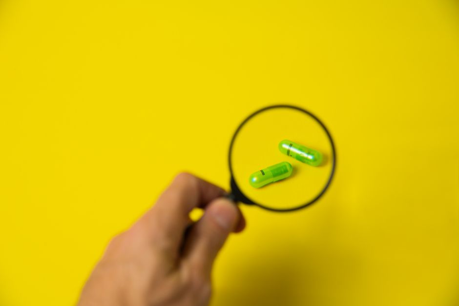 From above of crop unrecognizable male holding magnifying glass over green pills on yellow background in studio