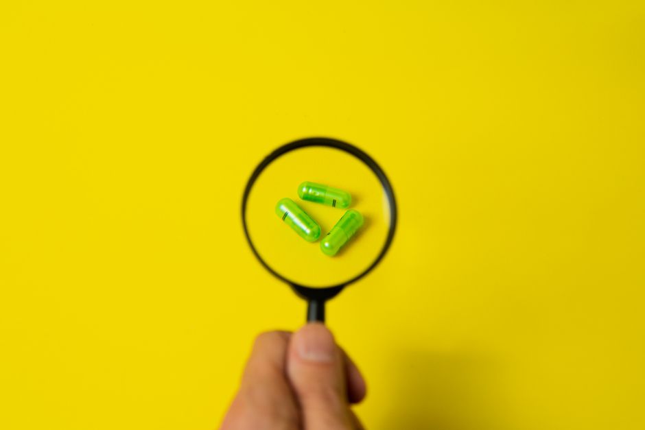 Top view of crop unrecognizable person holding magnifier over green pills placed on bright yellow surface in studio
